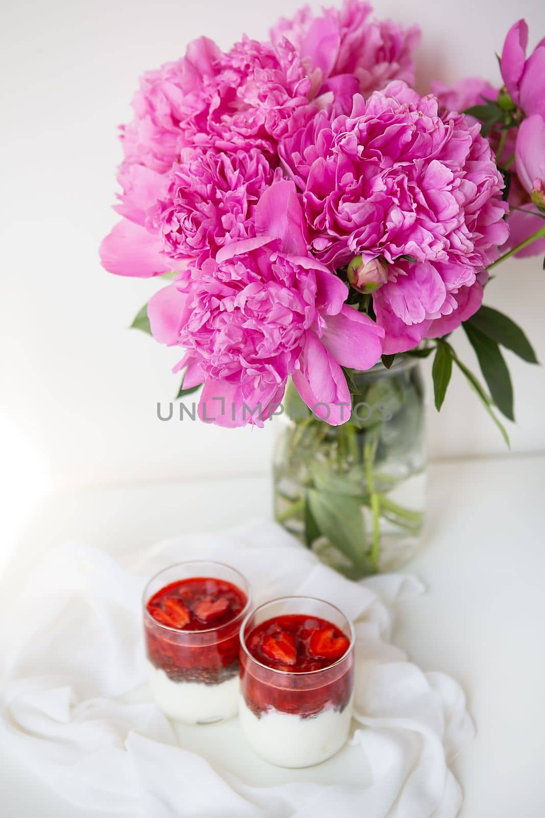 A beautiful bouquet of pink peonies in a vase on a white table stands along with a heart-shaped strawberry dessert. St. Valentine's Day, 8 March