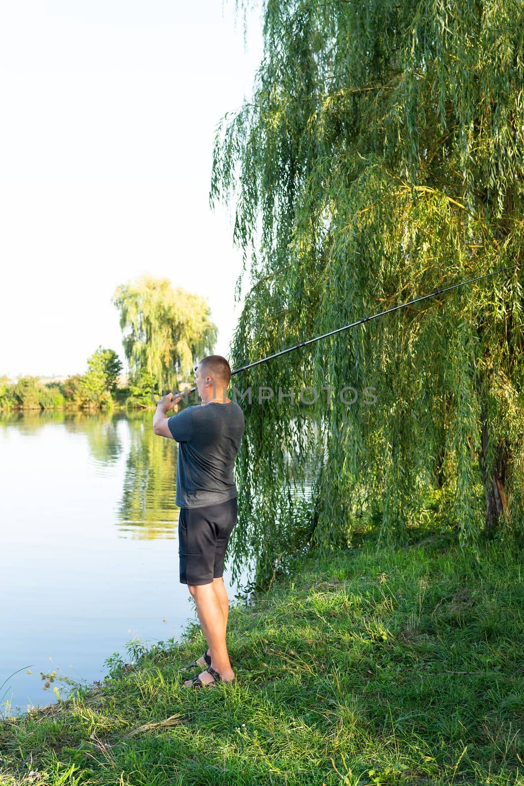 Fishing Reel. The guy throws a fishing rod against the backdrop of the lake. The concept of relaxation and hobby for the soul. by sfinks
