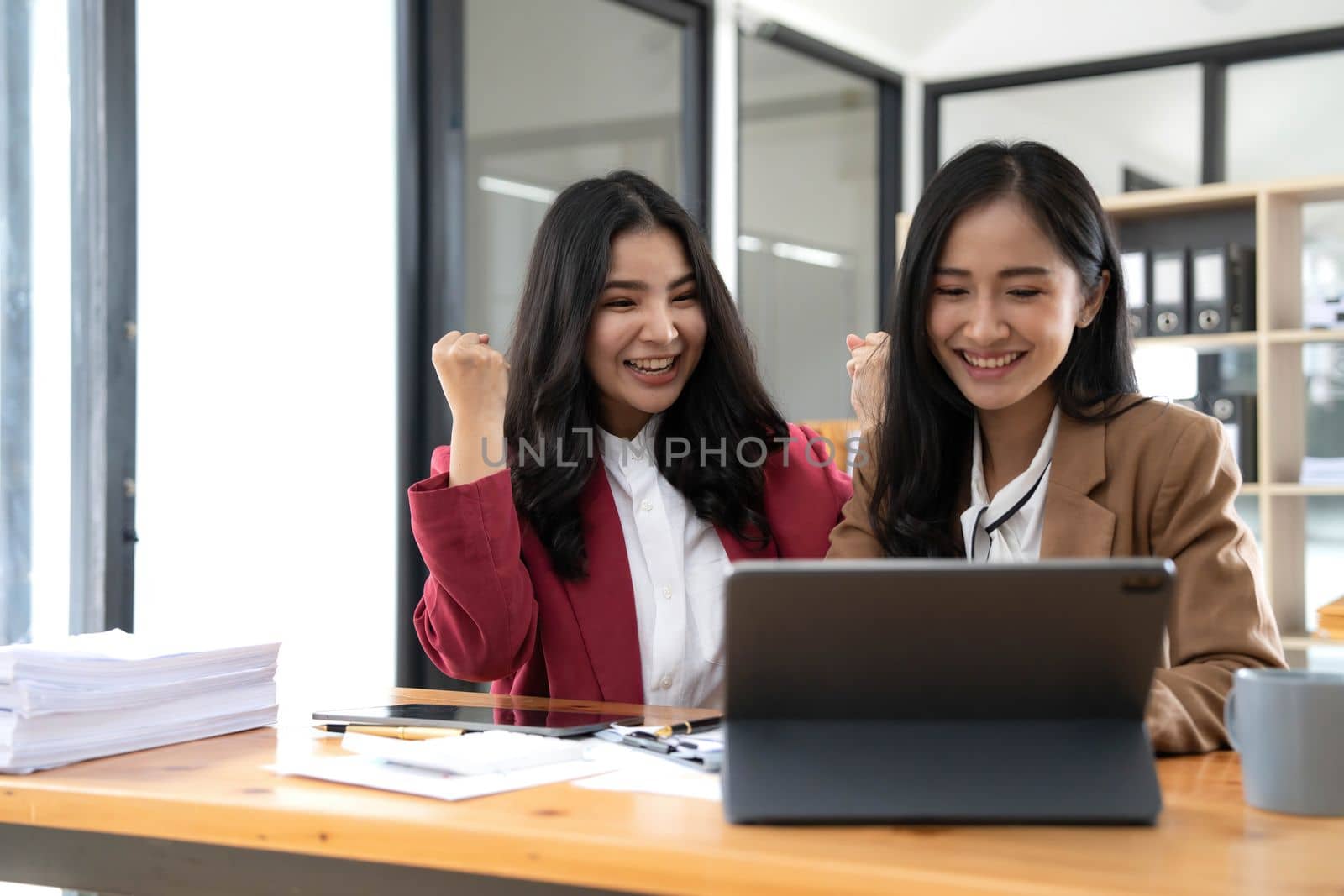 Two young Asian businesswomen show joyful expression of success at work smiling happily with a laptop computer in a modern office. by wichayada