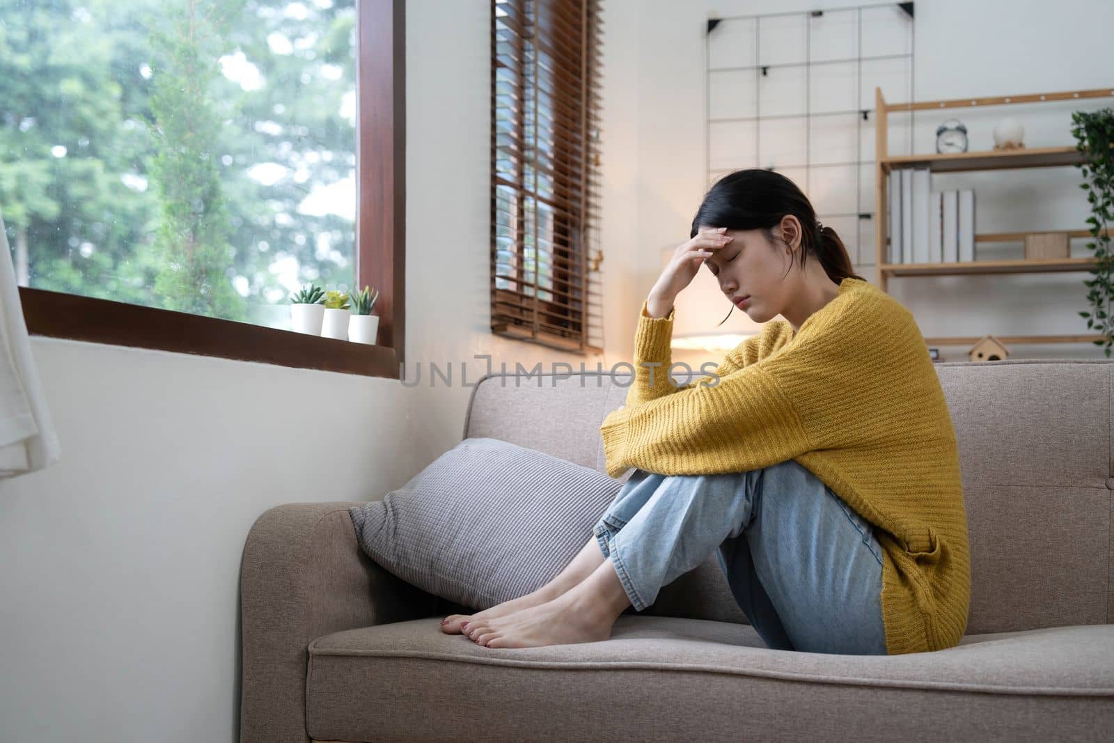 Hand on temples of young unhappy sadness Asian girl sitting on sofa. She is feeling not very good due to her sickness and having a headache. Seen from the front. Stress and sickness concept..