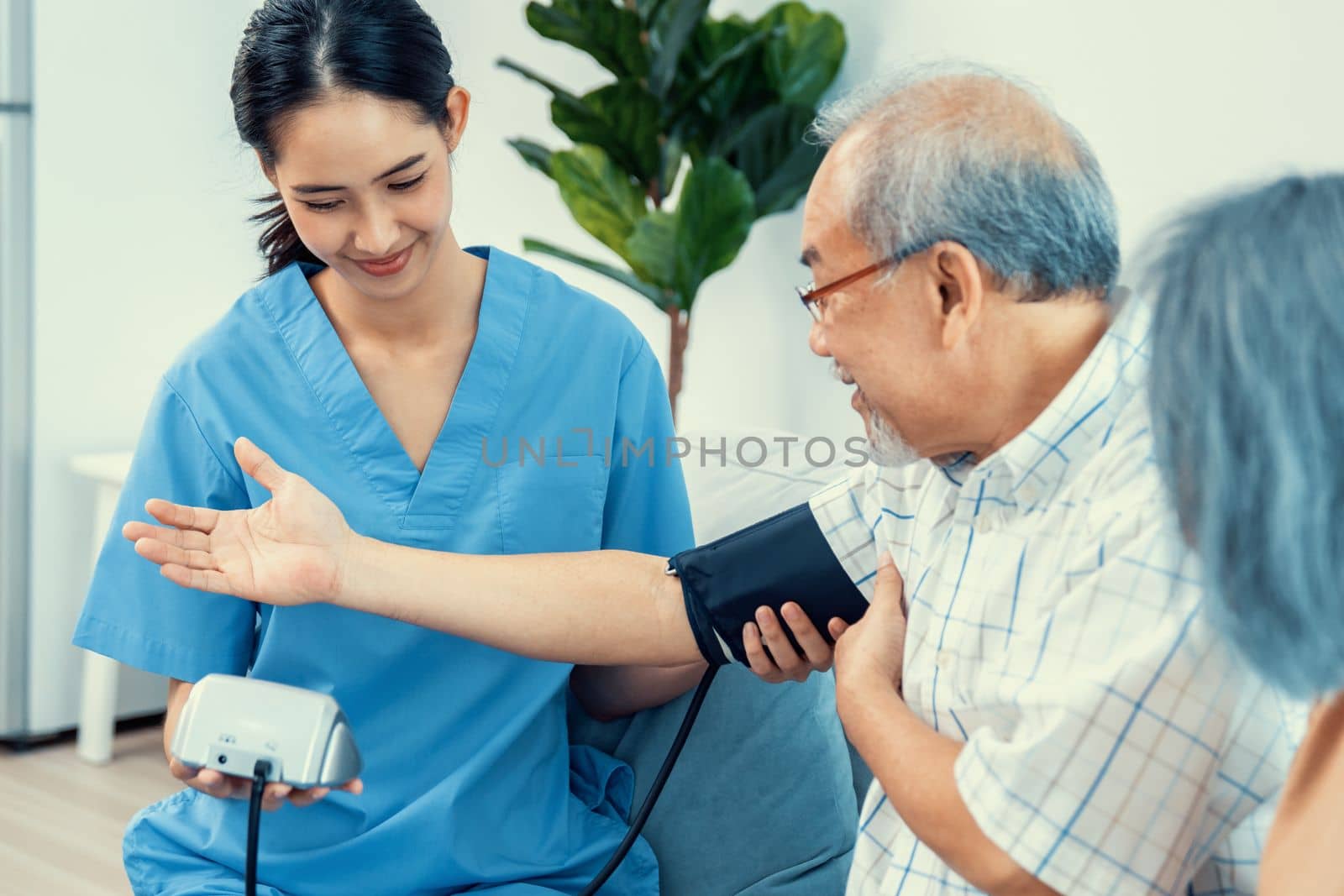 An elderly man having a blood pressure check by his personal caregiver with his wife sitting next to him in their home.