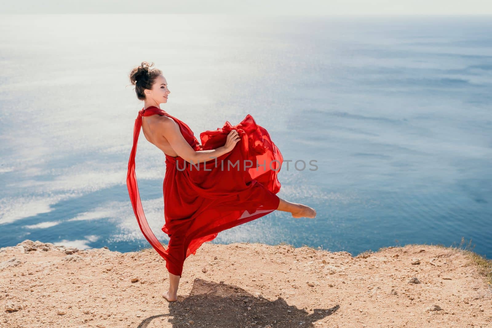 Woman in red dress on sea. Side view a Young beautiful sensual woman in a red long dress posing on a rock high above the sea on sunset. Girl on the nature on blue sky background. Fashion photo. by panophotograph