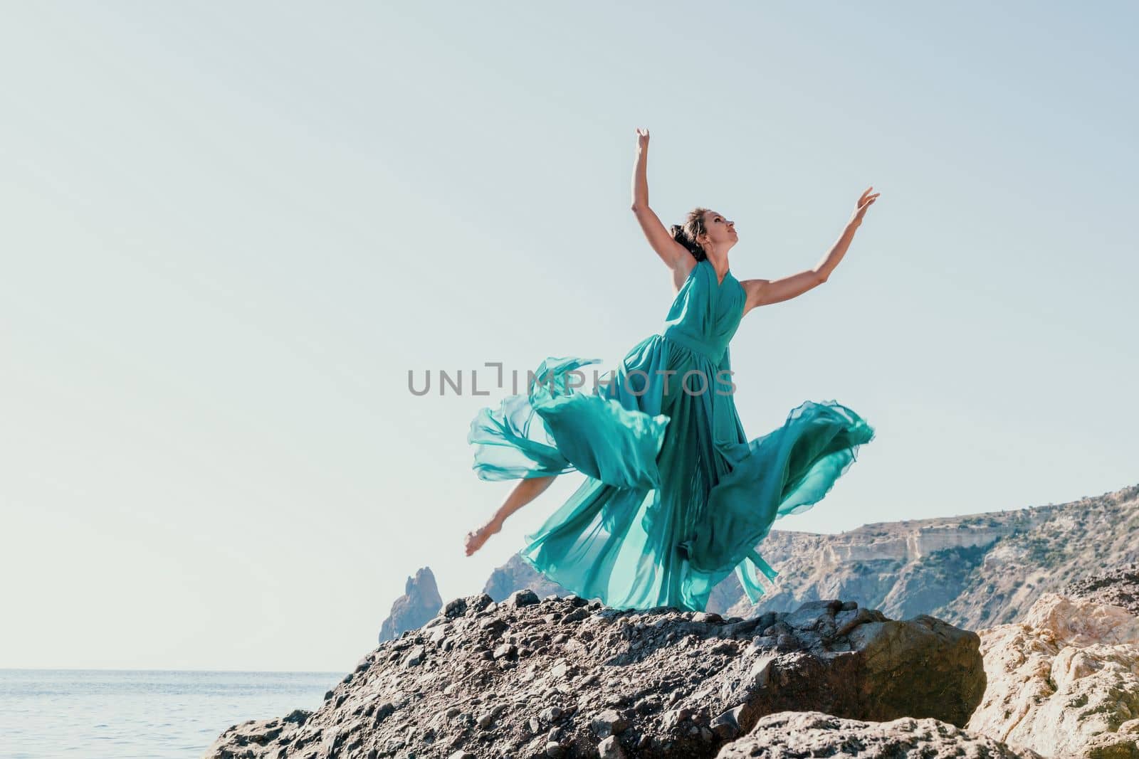 Side view a Young beautiful sensual woman in a red long dress posing on a rock high above the sea during sunrise. Girl on the nature on blue sky background. Fashion photo.