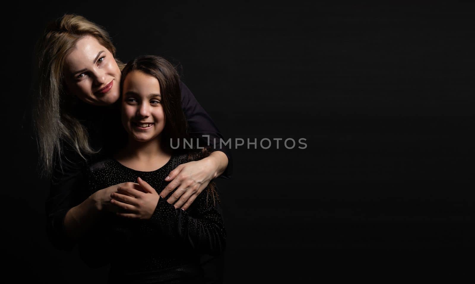 Young mother and her little daughter are looking at camera and smiling, sitting against black wall.
