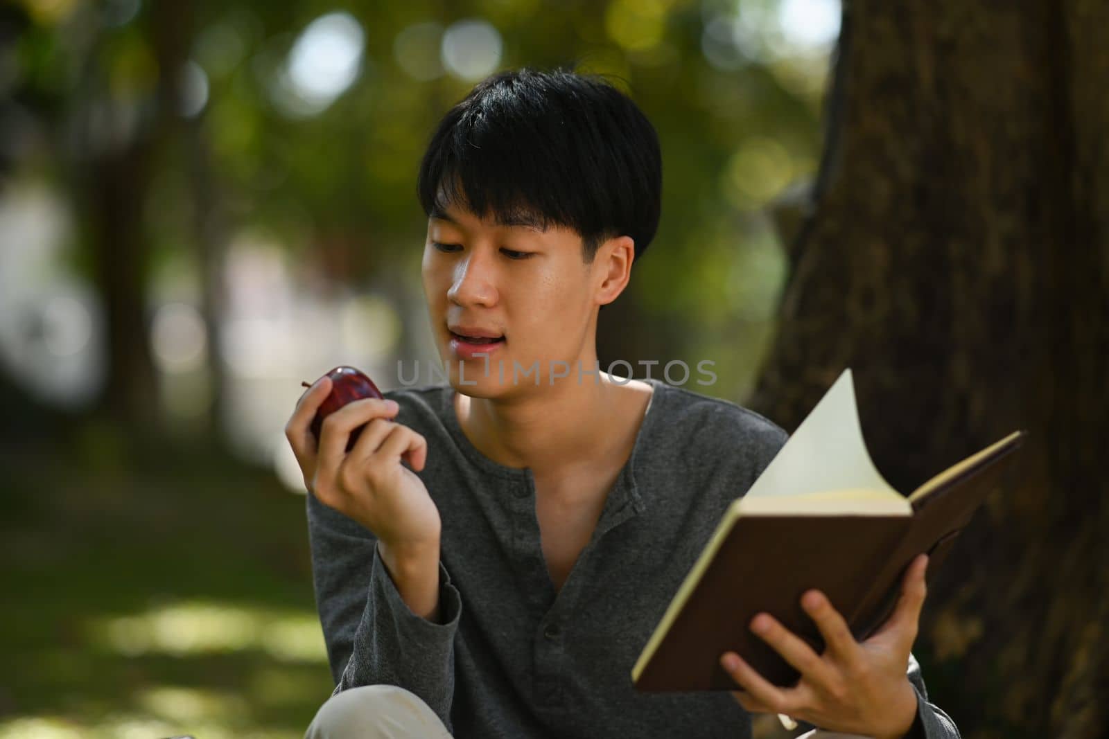Happy young asian man eating apple and reading book under tree at sunny beautiful garden background.