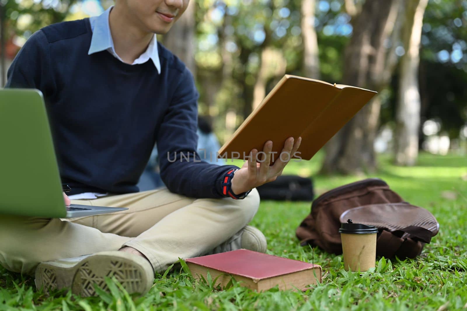 Cropped shot of male college student using laptop on green meadow in campus. Education, youth and technology concept by prathanchorruangsak