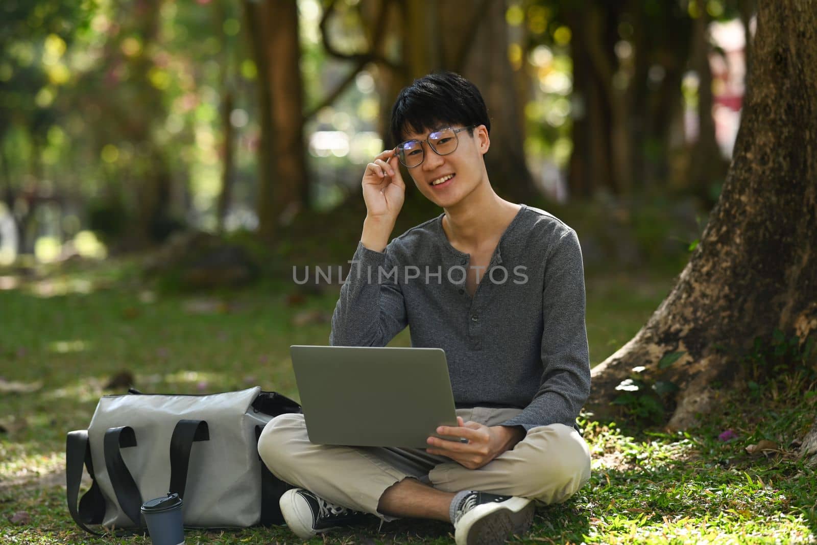 Pleasant asian man using laptop under a tree surrounded by beautiful nature. Education and lifestyle concept by prathanchorruangsak