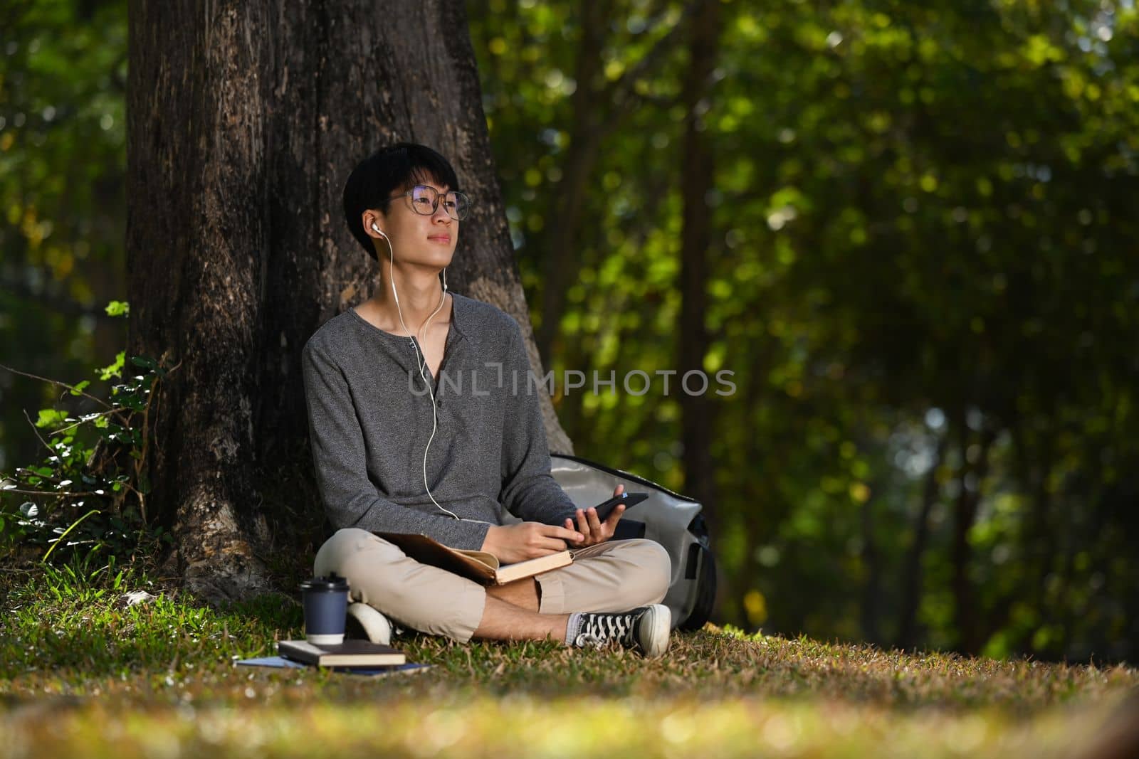Relaxed asian man enjoy listening music and reading book under the tree in the park.