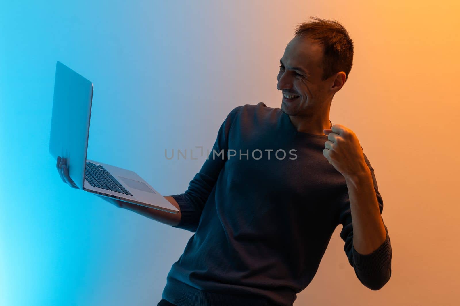 Emotional young man with laptop celebrating victory on color background.