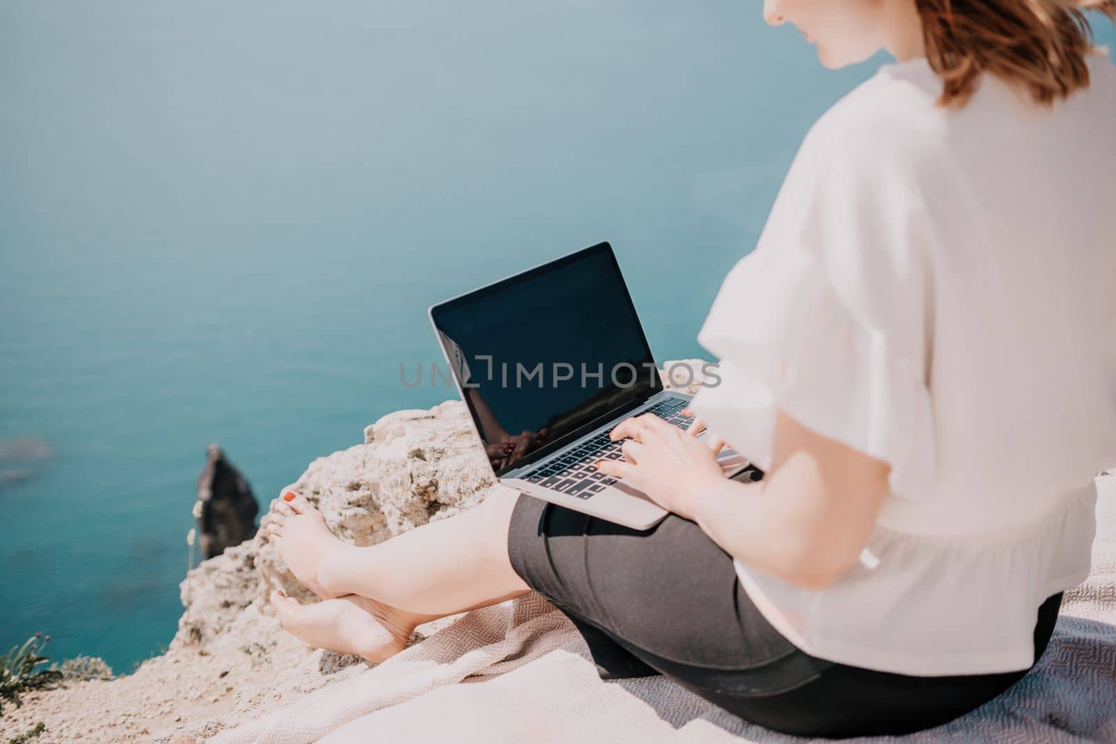 Successful business woman in yellow hat working on laptop by the sea. Pretty lady typing on computer at summer day outdoors. Freelance, travel and holidays concept.