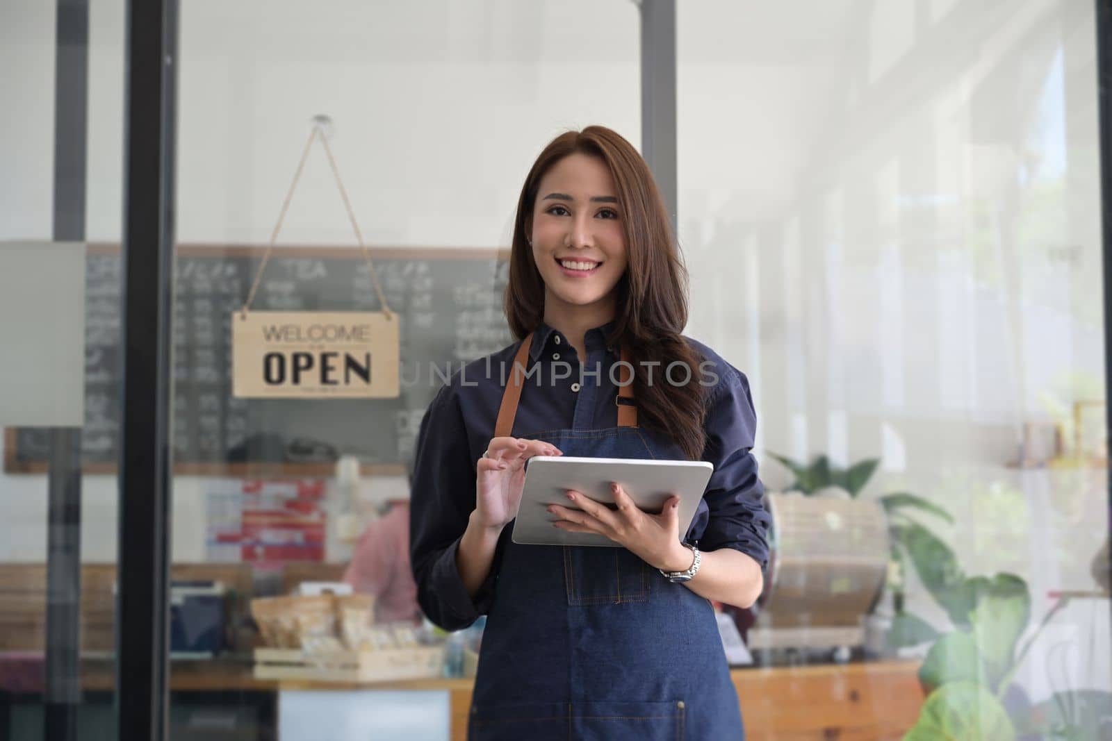 Young female entrepreneur in apron and using digital tablet while standing in her modern cafe.