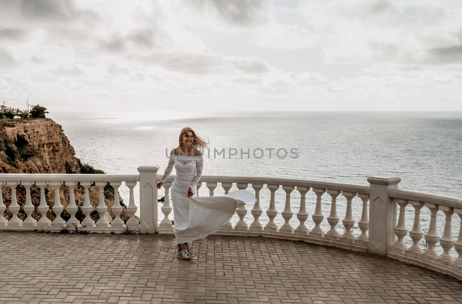Romantic bride, blond girl in white wedding dress with open shoulders posing on open terrace with backdrop of the sea and rocks. Stylish young woman standing on terrace and looking on ocean at sunset by panophotograph