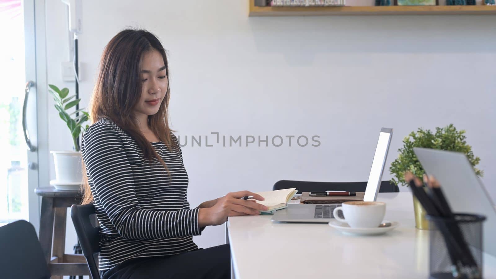 Young Asian woman reading a book and using laptop computer in cafe.