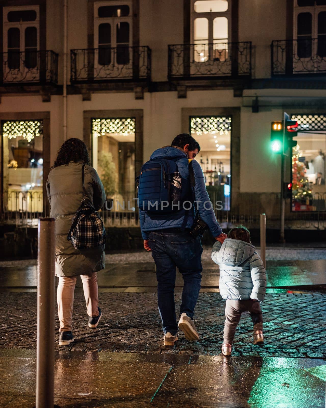 father holds hand of little daughter and family crossing road at night with the green traffic light.