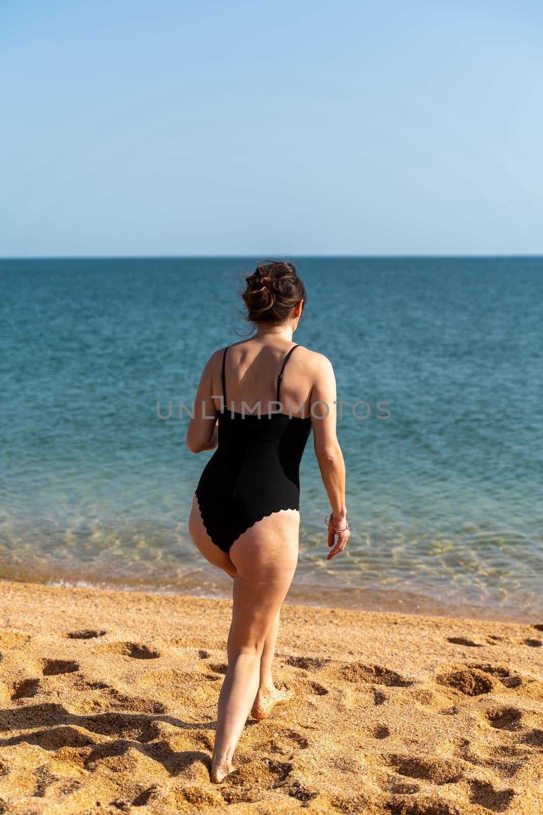 Woman sea swimsuit sand. A girl in full growth stands with her back and enters the sea in a black swimsuit. Alone on the beach on a sunny day