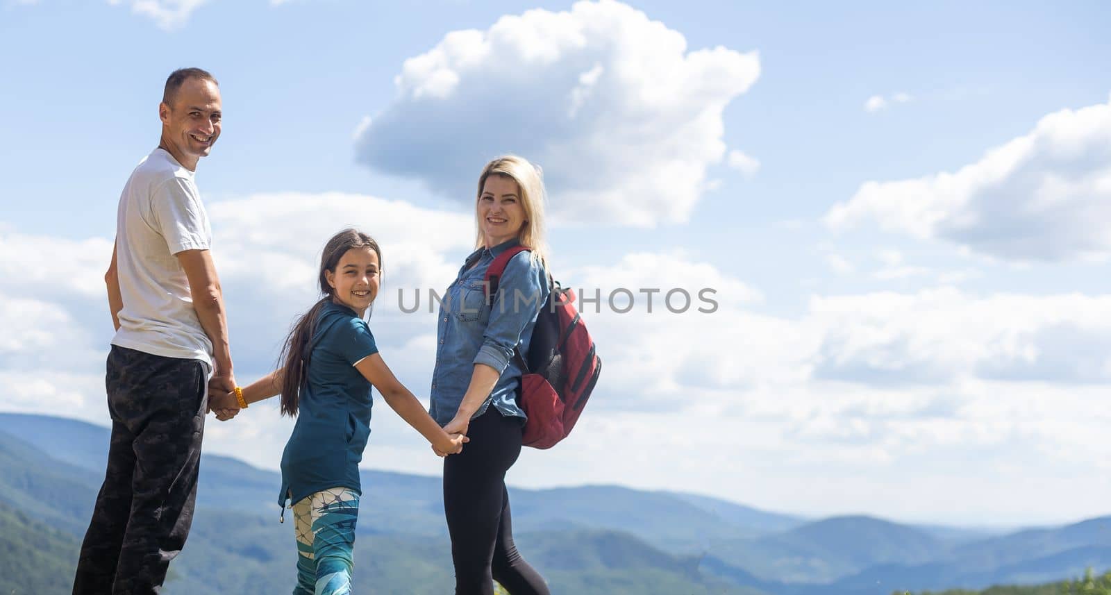 Mother, father hugging son, and daughter in summer mountains. Happy family smiling and standing on the green grass in the field at sunset. Kids with parents. Family weekend.