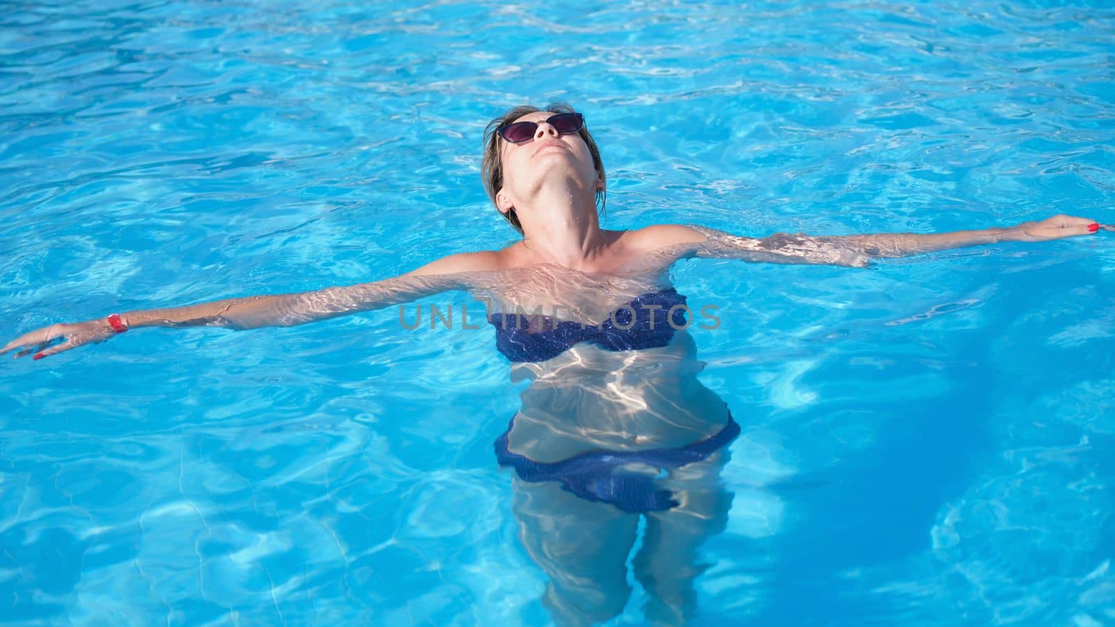 Glamorous woman in stylish sunglasses enjoys swimming in turquoise water in pool. Relaxing in pool of luxury resort