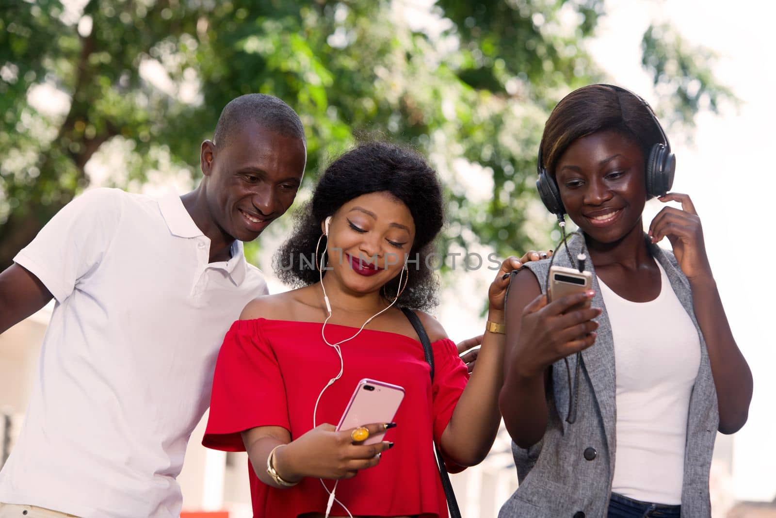 group of three happy young people with mobile phone takes picture and listens to music with headphone outside