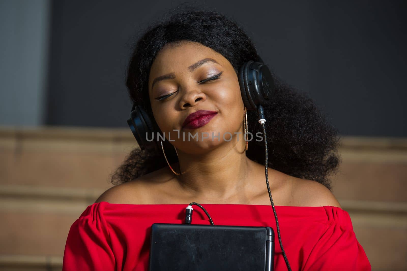 young girl sitting in red camisole with laptop listening to music using headphones with eyes closed while smiling.