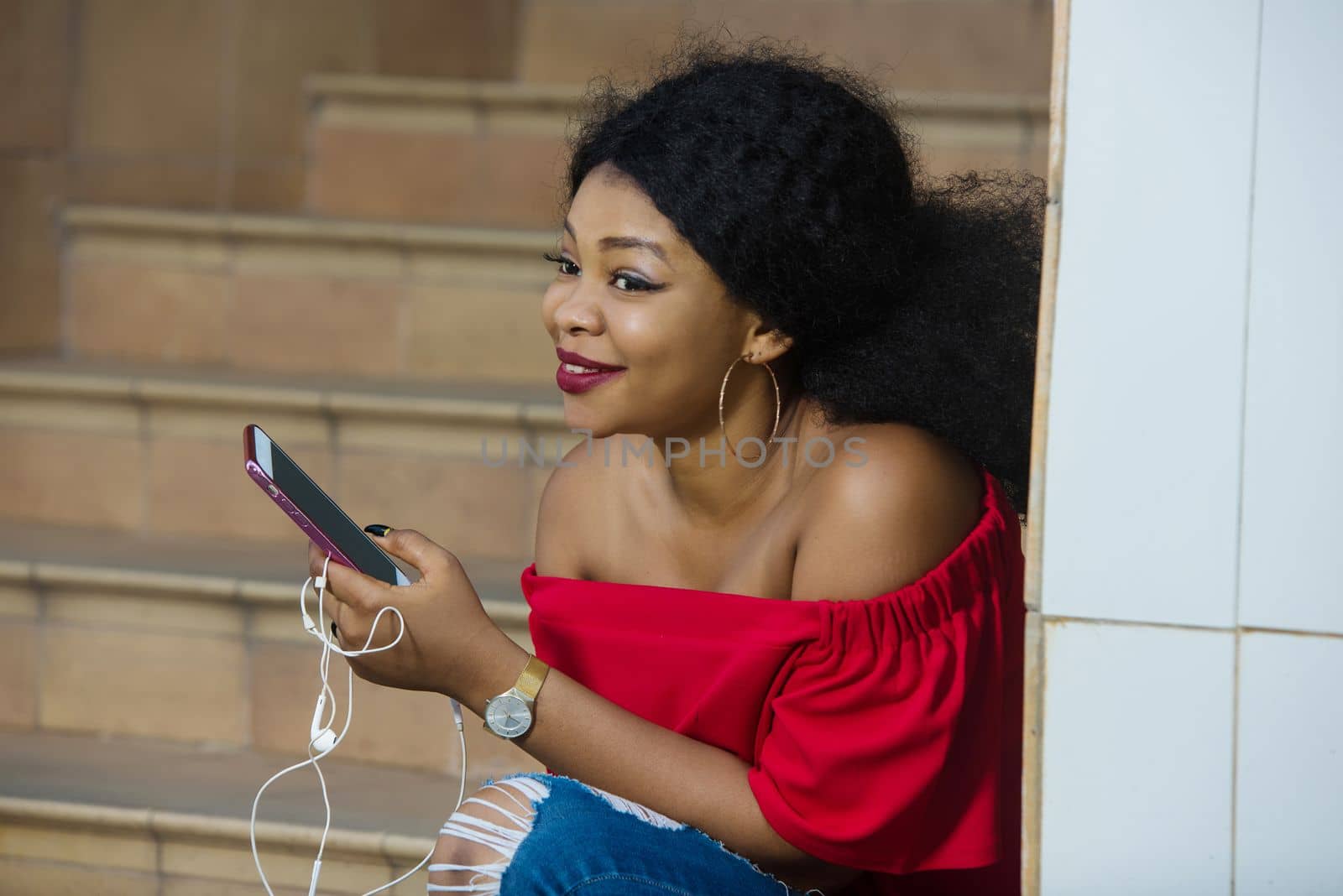 Funny african woman sitting on stairs listening to music with headphones and a smart phone outdoors.
