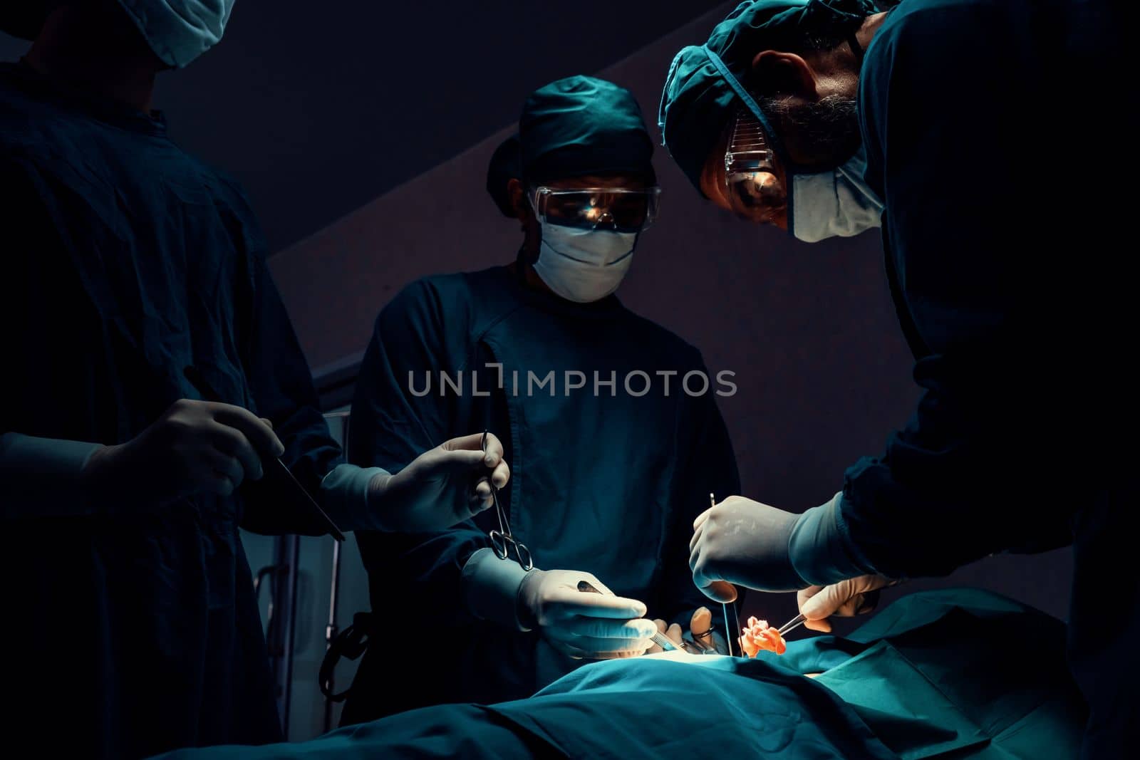 Surgical team performing surgery to patient in sterile operating room. In a surgery room lit by a lamp, a professional and confident surgical team provides medical care to an unconscious patient.