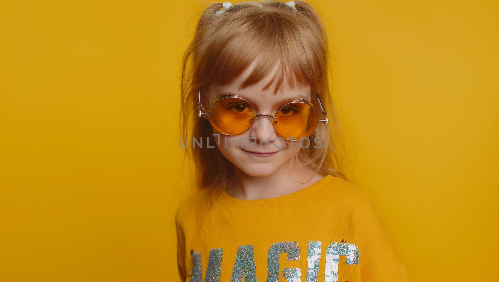 Smiling playful happy young preteen child girl kid walking slowly and looking at camera, flirting. Little charismatic lovely toddler children in orange sunglasses isolated on yellow wall background