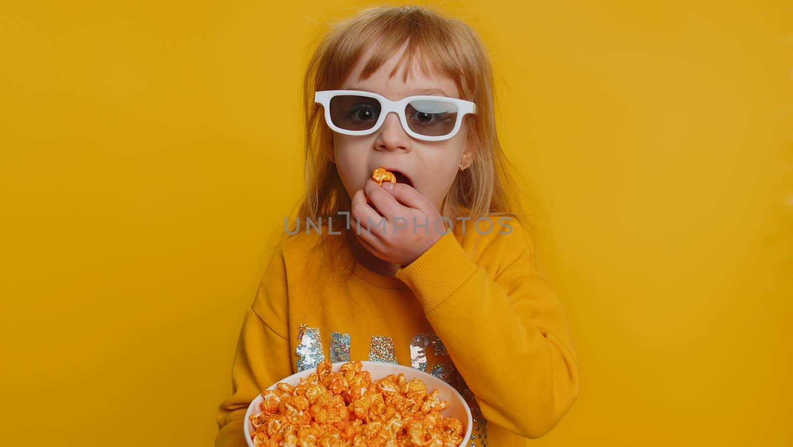 Excited young toddler school girl in 3D glasses eating popcorn, watching interesting tv serial, sport game, film, online social media movie content. Teen female child kid on studio yellow background