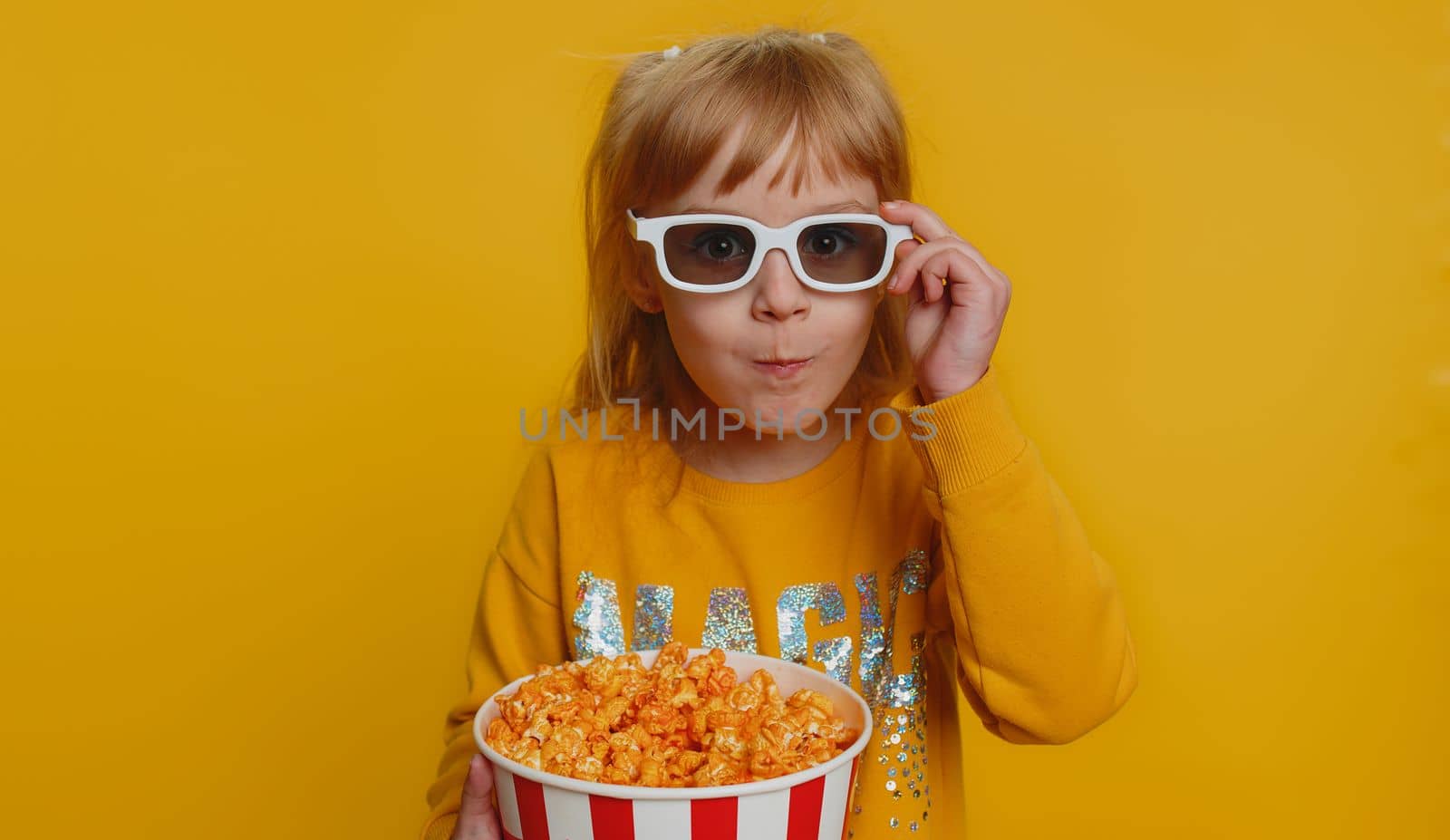 Excited young toddler school girl in 3D glasses eating popcorn, watching interesting tv serial, sport game, film, online social media movie content. Teen female child kid on studio yellow background