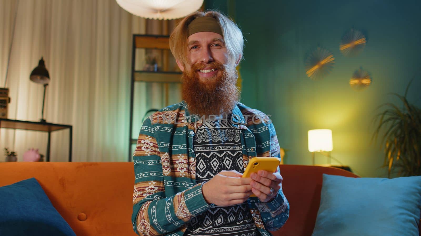 Hipster redhead bearded man sitting on sofa uses mobile phone smiles at night evening home. Young guy texting share messages content on smartphone social media applications online watching relax movie