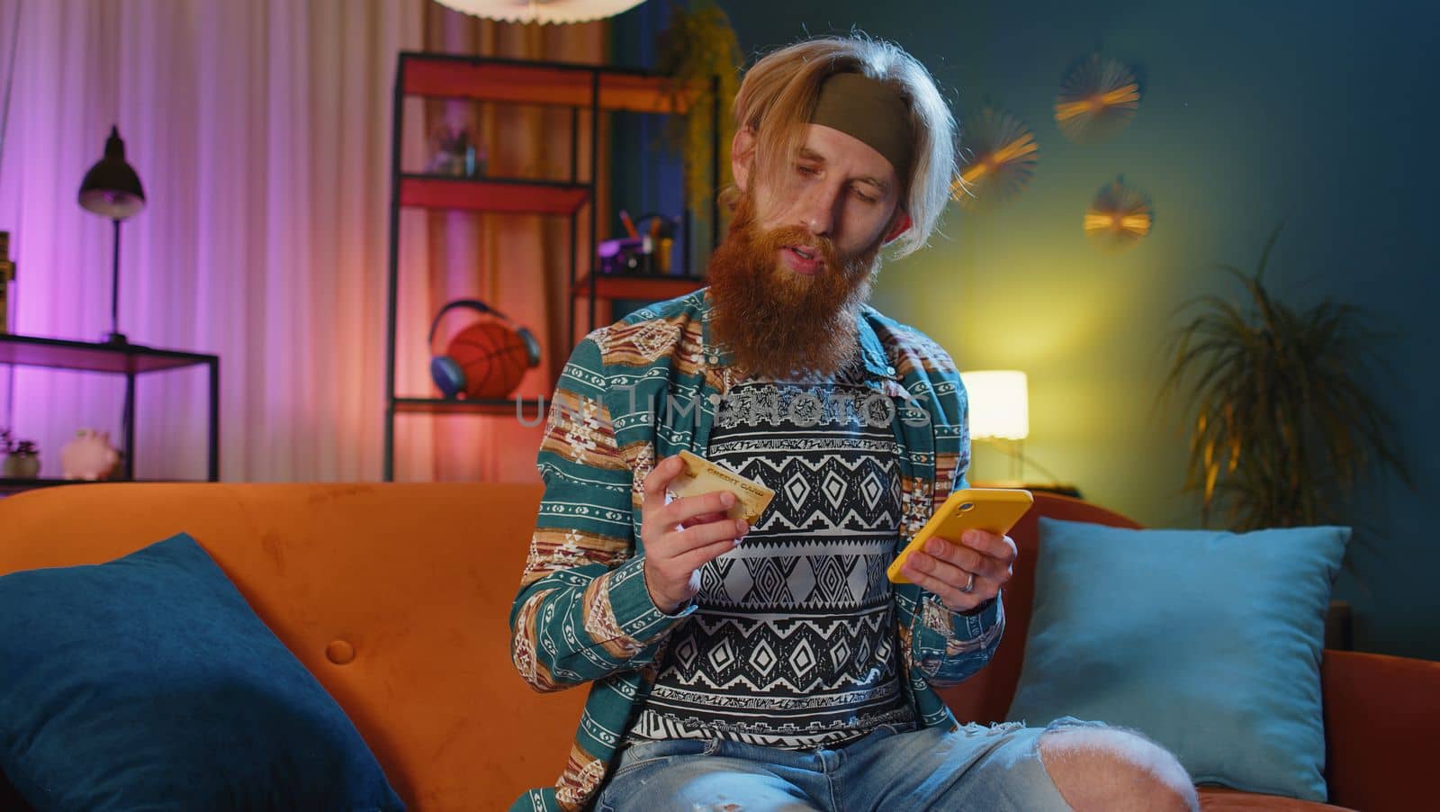 Bearded hippie man using credit bank card and smartphone while transferring money, purchases online shopping, order food delivery at home apartment indoors. Young redhead guy in evening room on couch