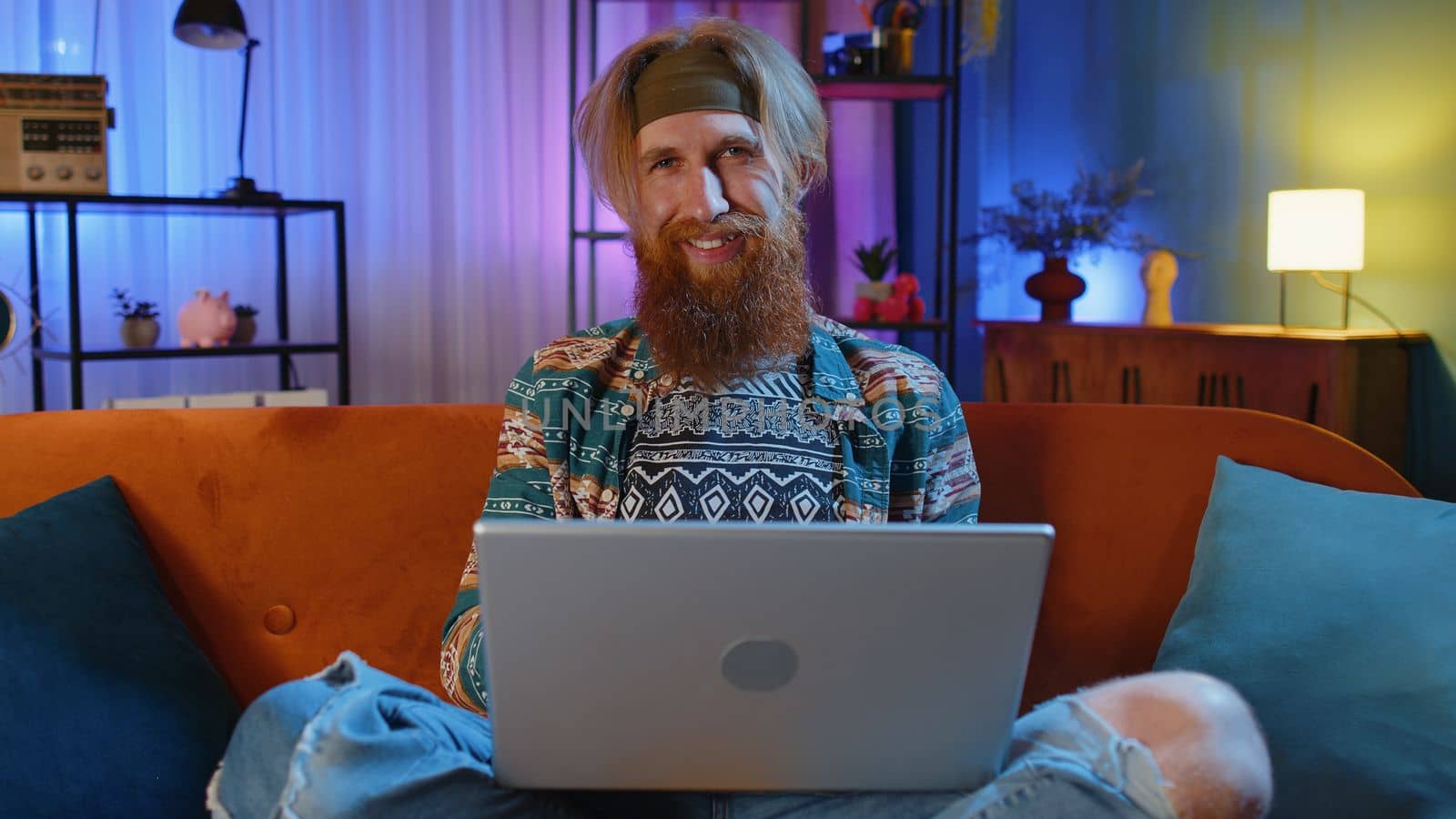 Handsome hippie bearded man freelancer at home night room sitting on couch, opens laptop start working. Young guy works on notebook, sends messages, makes online purchases, watching movies, working