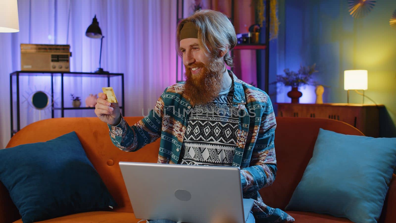 Bearded redhead man using credit bank card and laptop computer while transferring money, purchases online shopping, order food delivery at evening home. Young hippie guy on couch at home evening room