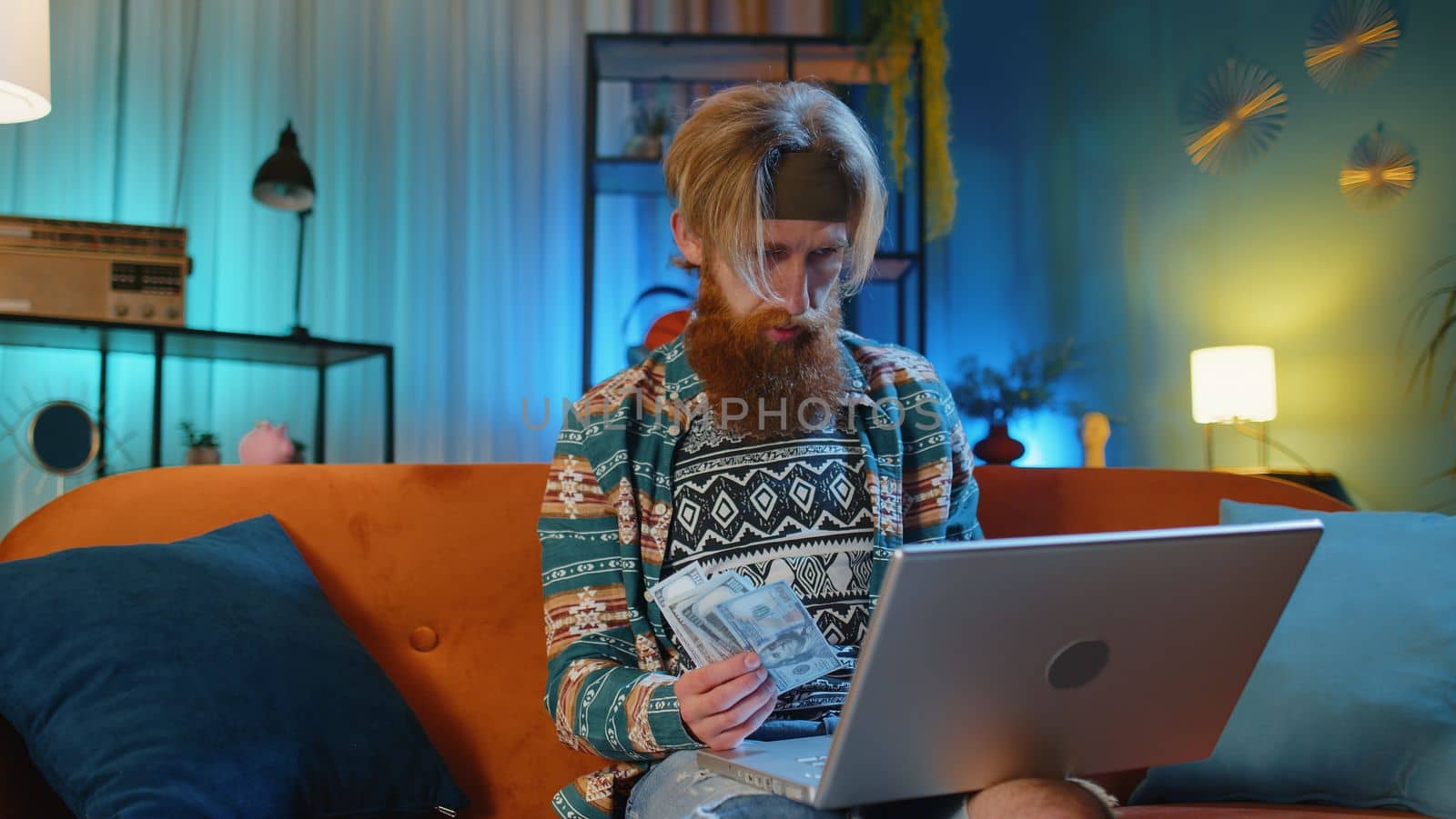 Planning budget. Rich happy hippie man counting money cash use laptop calculator, calculate domestic bills at home. Young guy satisfied of income earnings, win, saves money for planned vacation, gifts