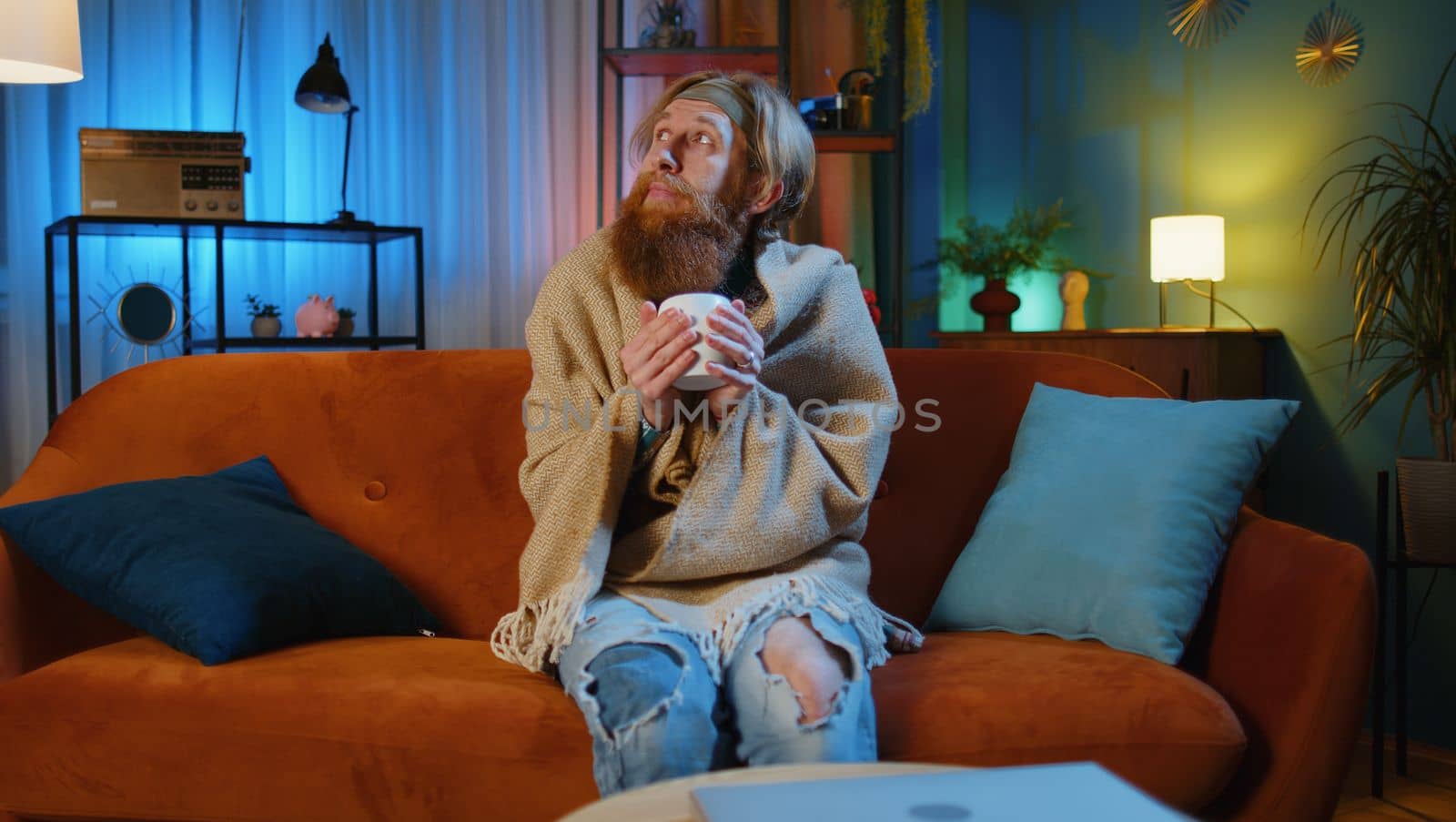 Sick man wrapped in plaid sit alone shivering from cold on sofa drinking hot tea in unheated apartment without heating due debt. Unhealthy young guy feeling discomfort try to warm up at evening home