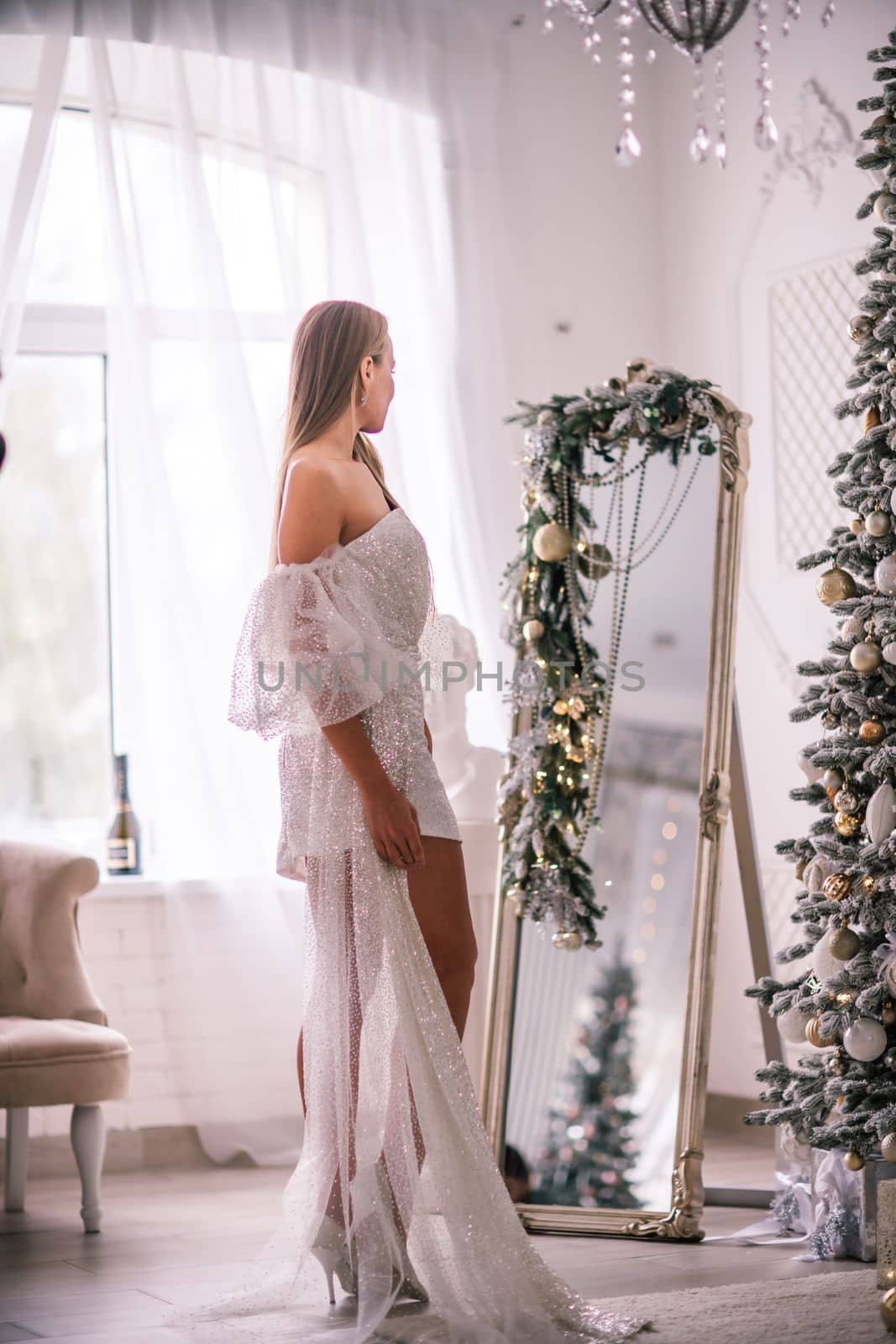 The blonde in the Christmas room. A beautiful blonde woman in a shiny light short dress with a train stands in a beautiful bright room decorated with a festive interior with a Christmas tree