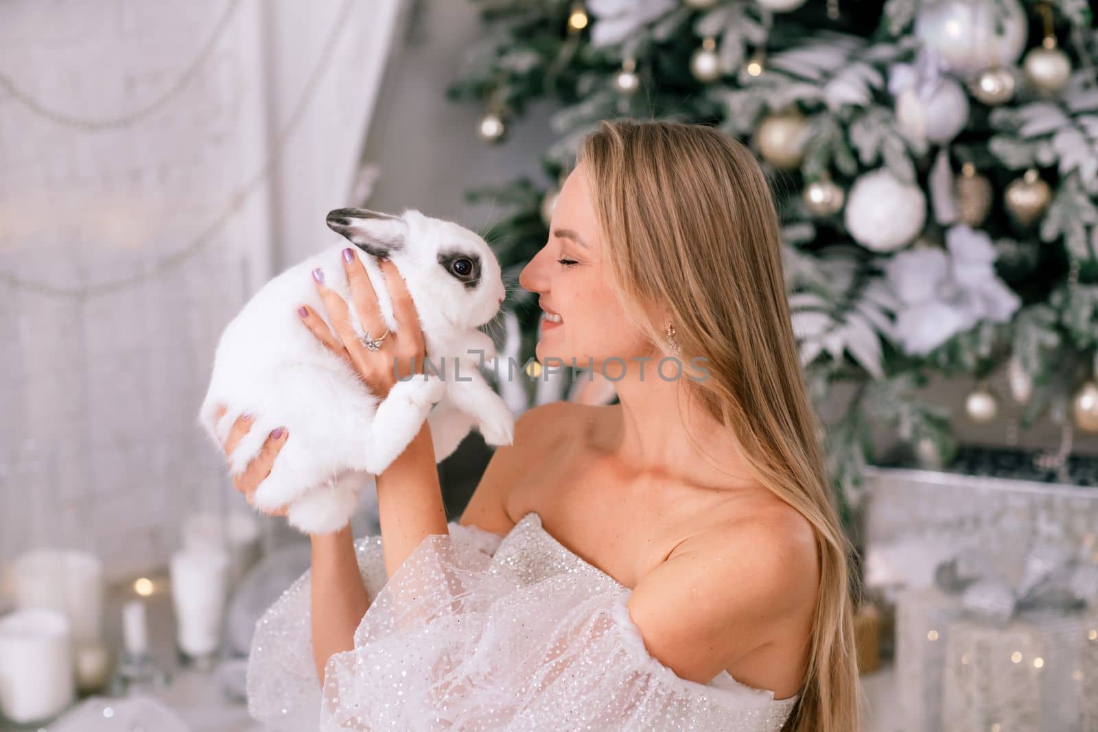 Woman holding a white rabbit symbol of the year 2023. Close-up of a beautiful young blonde woman holding a rabbit in a sparkly dress. She sits in a Christmas decorated room.