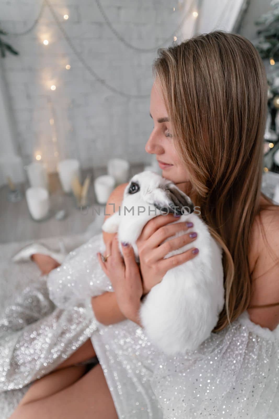 Woman holding a white rabbit symbol of the year 2023. Close-up of a beautiful young blonde woman holding a rabbit in a sparkly dress. She sits in a Christmas decorated room.