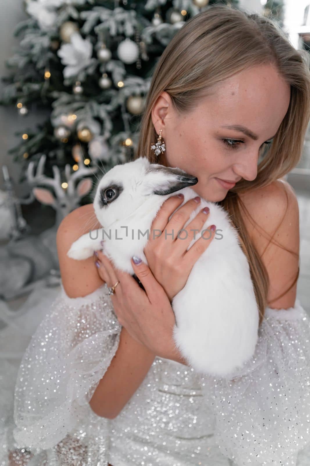 Woman holding a white rabbit symbol of the year 2023. Close-up of a beautiful young blonde woman holding a rabbit in a sparkly dress. She sits in a Christmas decorated room by Matiunina