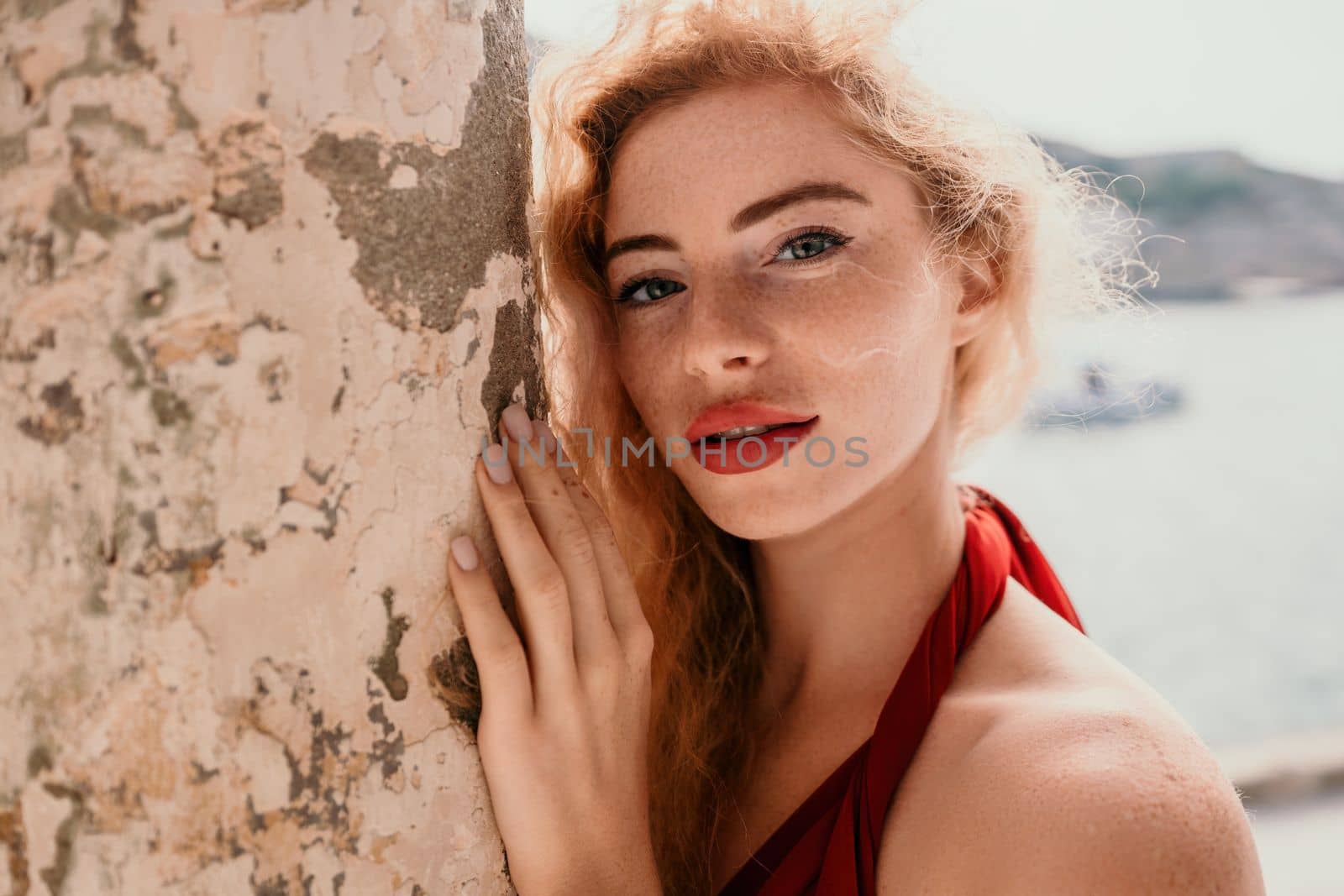 Close up shot of beautiful young caucasian woman with curly blond hair and freckles looking at camera and smiling. Cute woman portrait in a pink long dress posing on a volcanic rock high above the sea