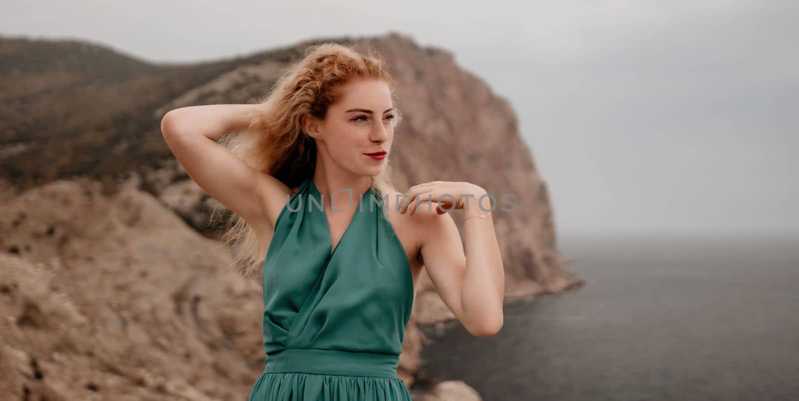 Redhead woman portrait. Curly redhead young caucasian woman with freckles looking at camera and smiling. Close up portrait cute woman in a mint long dress posing on a volcanic rock high above the sea by panophotograph