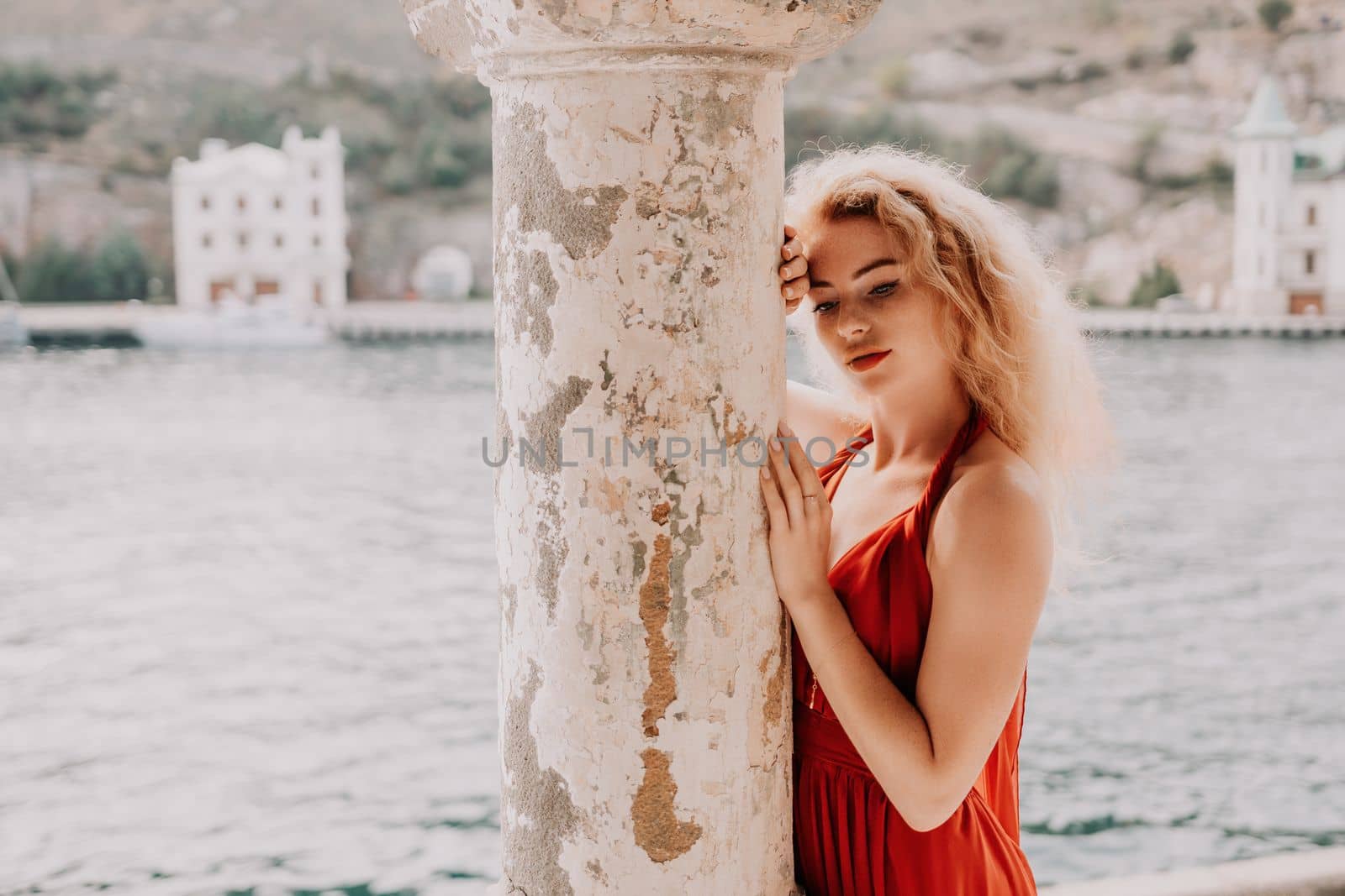 Close up shot of beautiful young caucasian woman with curly blond hair and freckles looking at camera and smiling. Cute woman portrait in a pink long dress posing on a volcanic rock high above the sea