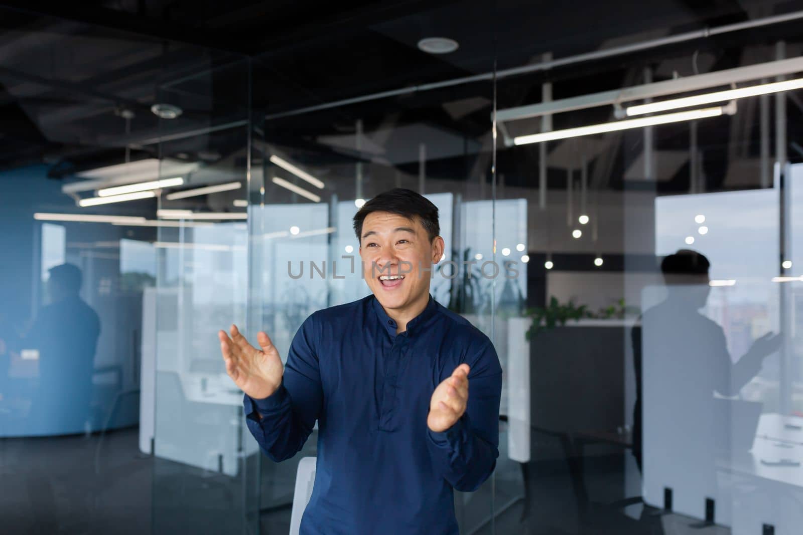 Young man Asian businessman, employee dancing in the office, celebrating victory, successful deal, monetary reward, promotion. He smiles, claps his hands.