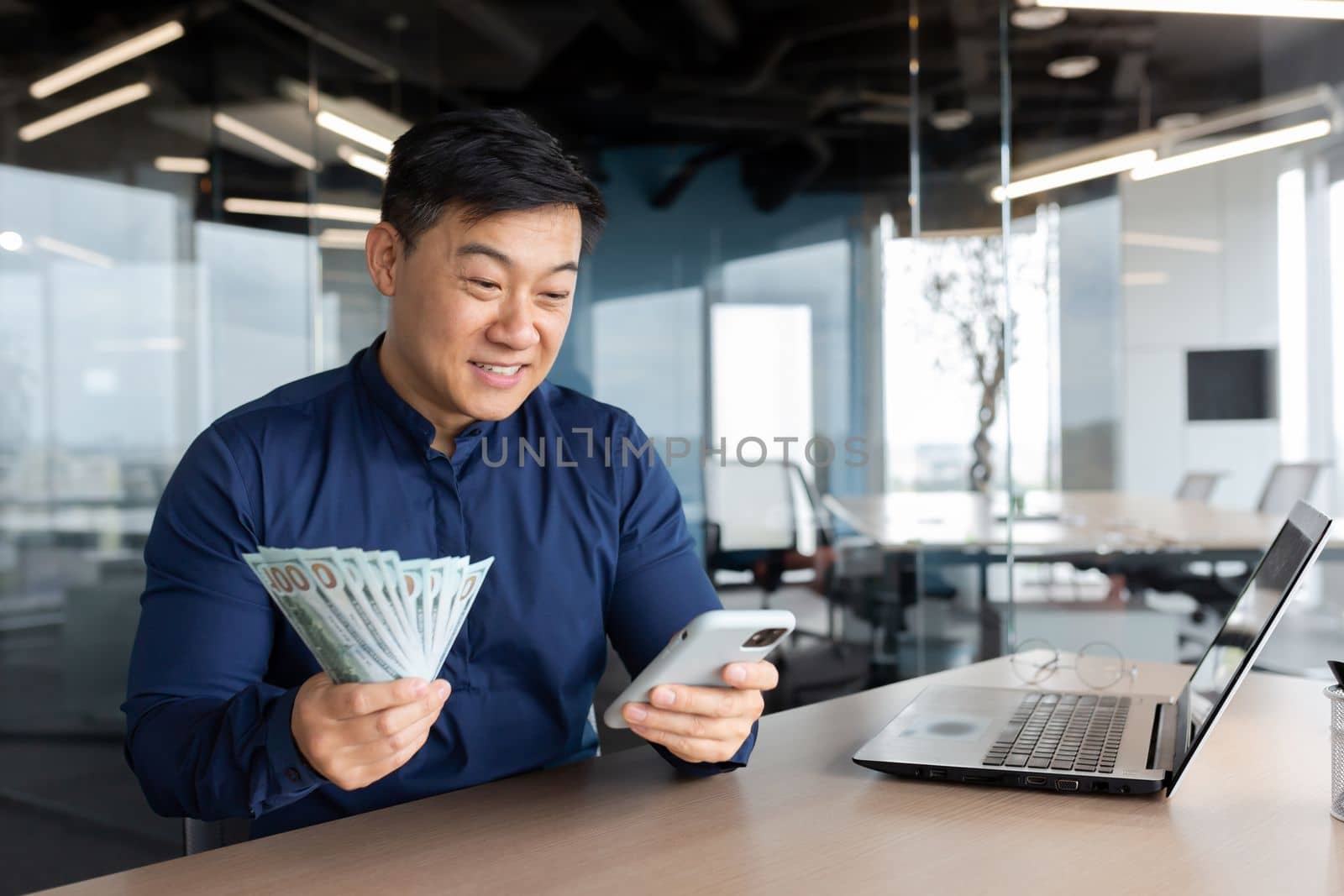 A young Asian businessman is sitting in the office at a table with a laptop. He is holding a phone and cash money. Plays online games, checks account, successful deal, investment.