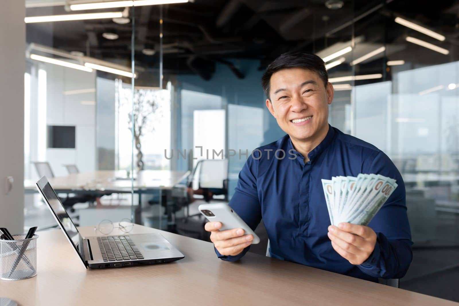 A young Asian man is sitting in the office. He is holding a phone and cash money. He looks at the camera, smiles.