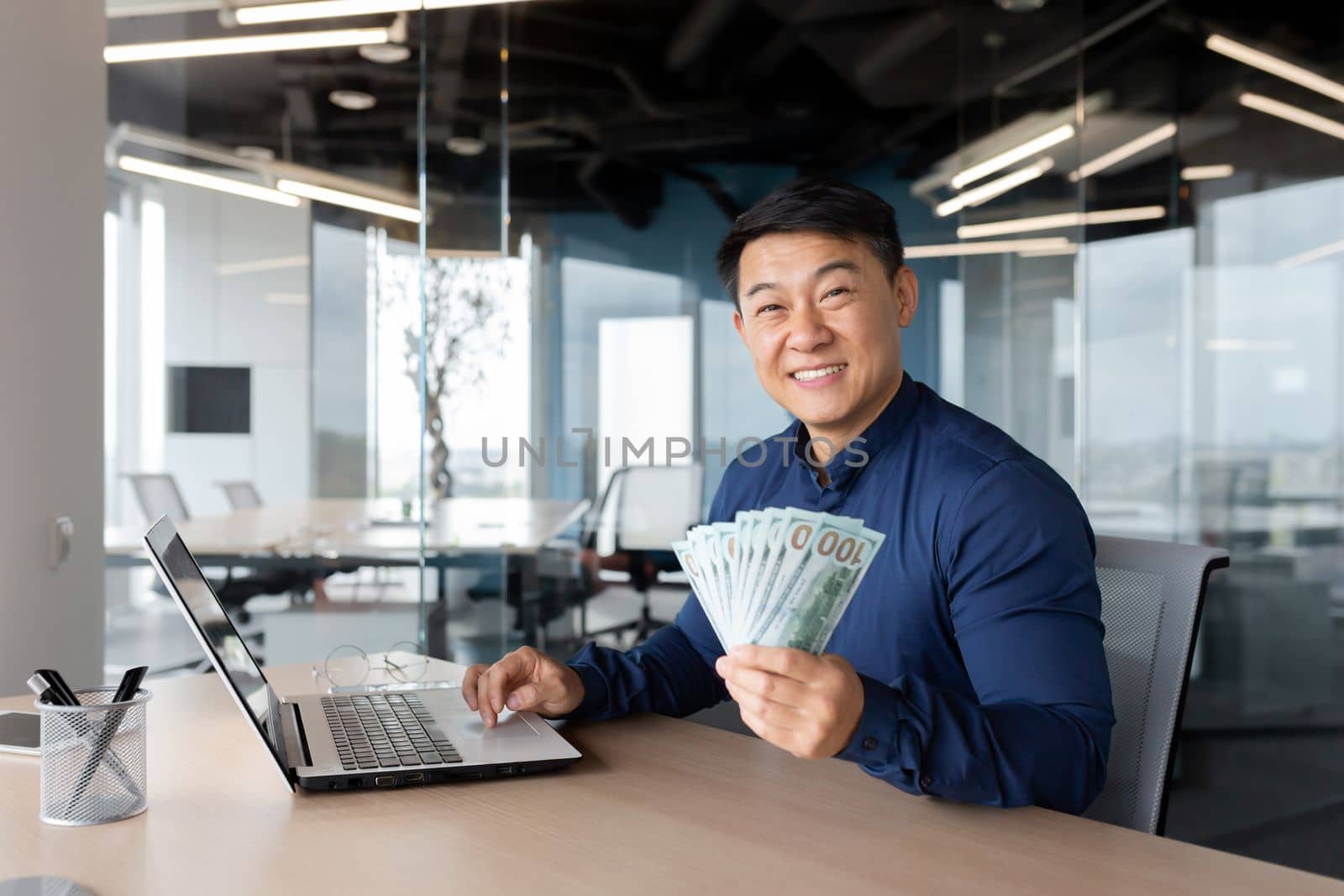A young Asian man sits in the office at a table with a laptop. He holds a phone and cash in his hands, points to the camera, smiles.