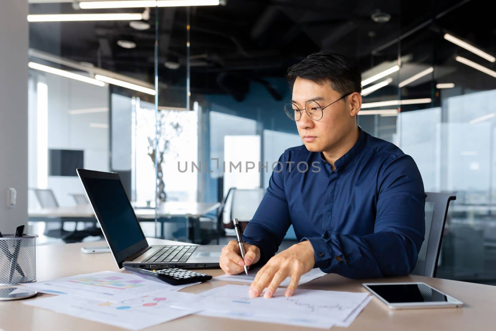 Concentrated young man businessman Asian works in the office at the table with laptop, writes documents, graphs, agreement, bills.