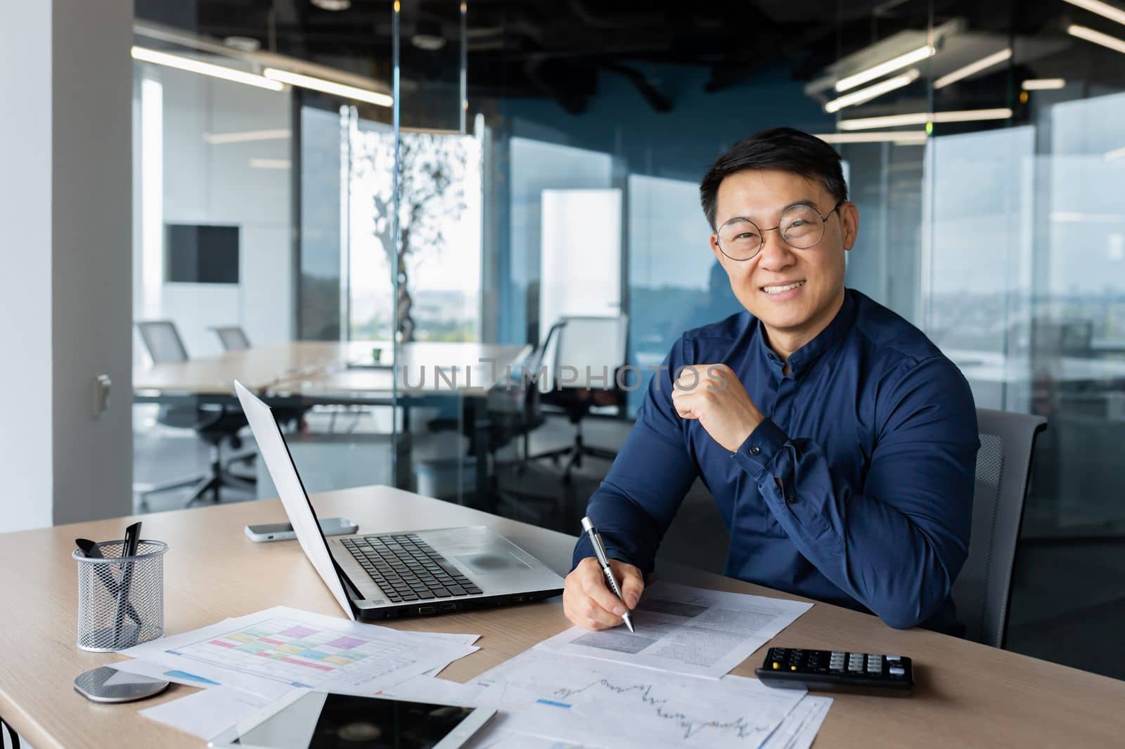 A successful young man businessman Asian works in the office at the table, works with documents, graphs, plan, accounts. Writes, looks at the camera, smiles.