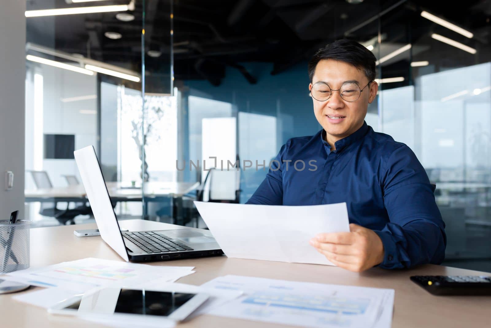 A young man, an Asian architect, designer, engineer, freelancer works in the office at a desk using a laptop. Holds documents, plans, drawings in his hands.