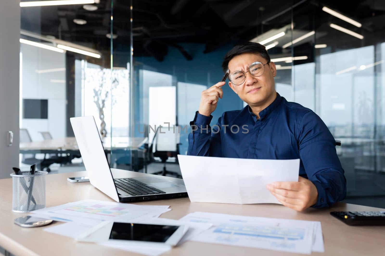 Serious young Asian architect, designer, engineer. Sitting at the desk in the office, holding documents and a pencil. The problem is working with projects, plans. Looking worriedly at the camera.