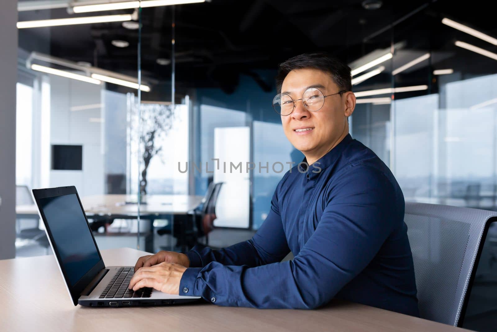 Portrait of a young male Asian student studying at a laptop. Sitting at the desk in the office, on campus, in the library. He studies online, remotely. He looks at the camera, smiles.